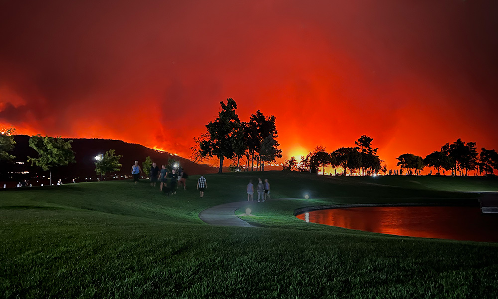 A landscape shot of a mountain on fire in the background, while people are in the foreground watching the fire from across the lake. The fire illuminates the night red and transforms it into shades of red and orange.